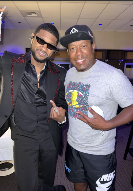 Nick Cooper, wearing a gray graphic T-shirt, black cap and black shorts, poses with singer Usher. Usher wears a black suit with red embellishments on the jacket and dark sunglasses as they smile for the camera indoors.